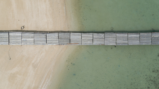 Aerial view of a wooden pier extending from a sandy beach into the clear turquoise waters of the tropical coast in Thailand. Aerial view of a wooden pier extending from a sandy beach into the clear turquoise waters of the tropical coast in Thailand.