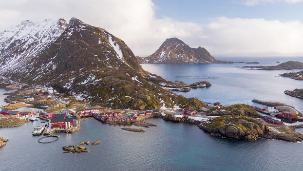 Aerial view of Lofoten with traditional red fishing cabins on rocky islands snow capped mountains and fish farming pens in the sea in Nordland Norway.