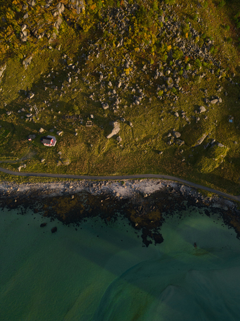 Aerial view of a small red house on a grassy slope with rocks next to a coastal road and turquoise water near Tangstad Leknes Nordland Norway. Aerial view of a small red house on a grassy slope with rocks next to a coastal road and turquoise water near Tangstad Leknes Nordland Norway.