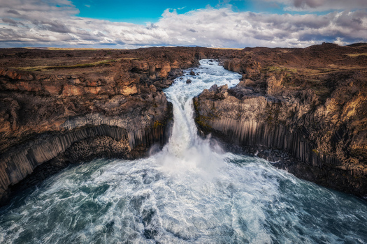 Aerial view of Aldeyjarfoss waterfall with powerful white water cascading over dark basalt columns under a cloudy sky in Iceland.