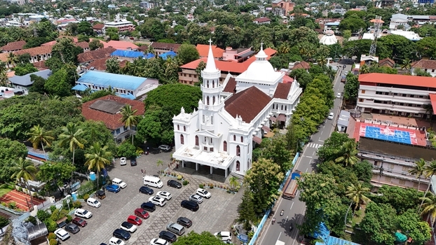 Aerial view of Mount Carmel Roman Catholic Cathedral with its white facade and tall spire surrounded by lush greenery parking area and urban streets in Alappuzha Kerala India. Aerial view of Mount Carmel Roman Catholic Cathedral with its white facade and tall spire surrounded by lush greenery parking area and urban streets in Alappuzha Kerala India.