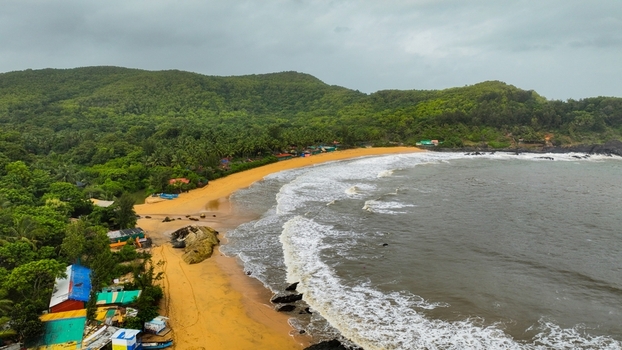 Aerial view of Gokarna Main Beach with golden sand crashing waves lush green hills and small coastal structures under a cloudy sky in Gokarna Karnataka India.