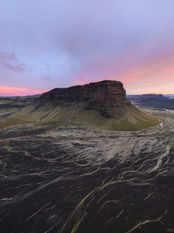 Aerial view of the majestic mountain rises dramatically from the dark textured plains under a sky painted with soft hues of pink and purple Skaftafell Skaftárhreppur Iceland. Aerial view of the majestic mountain rises dramatically from the dark textured plains under a sky painted with soft hues of pink and purple Skaftafell Skaftárhreppur Iceland.