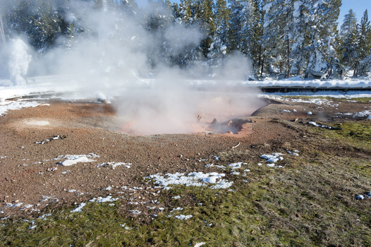 View of a steaming hot spring with orange mineral deposits surrounded by snow covered ground and pine trees in Yellowstone National Park Wyoming United States. View of a steaming hot spring with orange mineral deposits surrounded by snow covered ground and pine trees in Yellowstone National Park Wyoming United States.