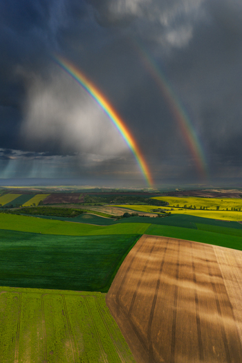 Aerial view of a double rainbow arching over vibrant agricultural fields with green and brown patterns under a dramatic stormy sky in Nasedlovice South Moravian Region Czechia.