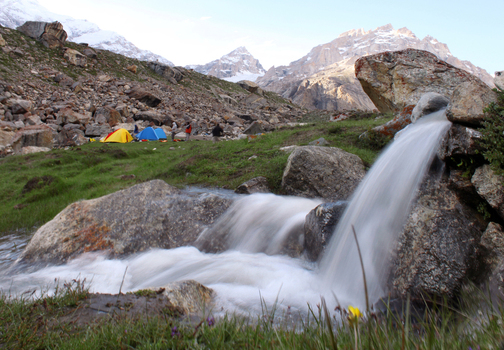 View of the Dalsanpa camping site on the K2 trek with a small stream flowing over rocks in the foreground and colorful tents in the rugged landscape of Hushe Pakistan.