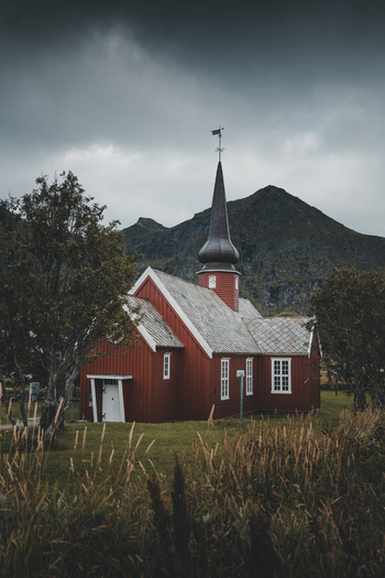 View of the historic red wooden Flakstad Church with its distinctive onion shaped spire set against dramatic mountains under a moody cloudy sky in Ramberg Nordland Norway. View of the historic red wooden Flakstad Church with its distinctive onion shaped spire set against dramatic mountains under a moody cloudy sky in Ramberg Nordland Norway.