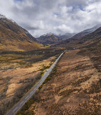 Aerial view of the A82 road winding through the dramatic Glen Coe valley surrounded by snow capped mountains and brown moorland under a cloudy sky in Scotland United Kingdom. Aerial view of the A82 road winding through the dramatic Glen Coe valley surrounded by snow capped mountains and brown moorland under a cloudy sky in Scotland United Kingdom.