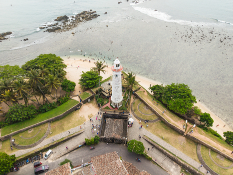 Aerial view of the historic Galle Lighthouse and stone fortifications of Galle Fort with people on the beach and palm trees in Galle Southern Province Sri Lanka. Aerial view of the historic Galle Lighthouse and stone fortifications of Galle Fort with people on the beach and palm trees in Galle Southern Province Sri Lanka.