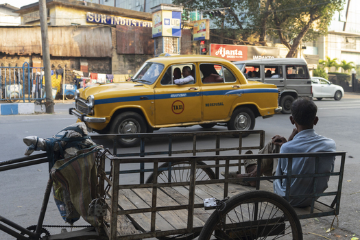 View of a classic yellow taxi speeds by a weathered handcart in Kolkata s vibrant streets blending old and new amidst the bustling city life Kolkata West Bengal India. View of a classic yellow taxi speeds by a weathered handcart in Kolkata s vibrant streets blending old and new amidst the bustling city life Kolkata West Bengal India.