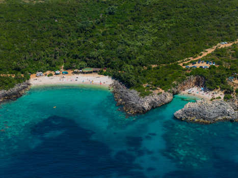 Aerial view of Aquarium Beach featuring turquoise waters rocky cliffs and lush green hills with people on the sand in Jala Vlore County Albania. Aerial view of Aquarium Beach featuring turquoise waters rocky cliffs and lush green hills with people on the sand in Jala Vlore County Albania.