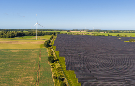Aerial view of a large solar farm with rows of photovoltaic panels next to a wind turbine in a rural landscape with green fields under a clear blue sky in Pomeranian Voivodeship Poland. Aerial view of a large solar farm with rows of photovoltaic panels next to a wind turbine in a rural landscape with green fields under a clear blue sky in Pomeranian Voivodeship Poland.