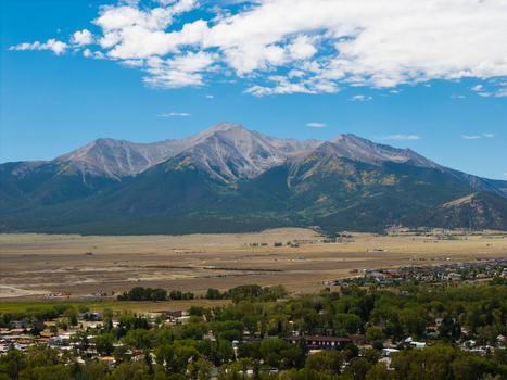 Aerial view of the Collegiate Peaks mountain range rising above the town of Buena Vista with green trees and open plains under a bright blue sky in Buena Vista Colorado United States.
