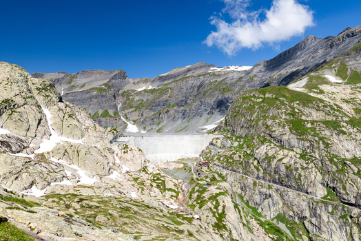 View of the Emosson Dam surrounded by rugged alpine mountains rocky slopes with patches of snow and a clear blue sky in Le Chatelard Fribourg Switzerland. View of the Emosson Dam surrounded by rugged alpine mountains rocky slopes with patches of snow and a clear blue sky in Le Chatelard Fribourg Switzerland.