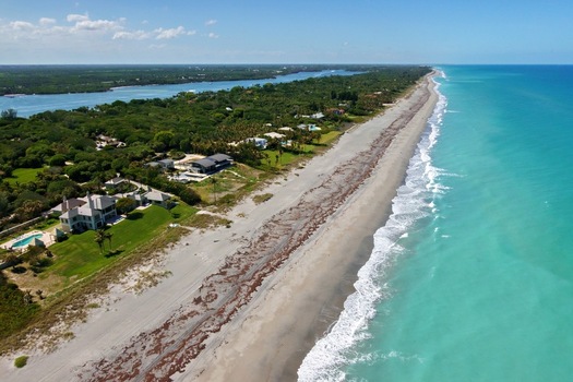 Aerial view of the turquoise coastline with luxury beachfront estates lush tropical vegetation and a wide sandy beach under a clear blue sky in Jupiter Florida United States. Aerial view of the turquoise coastline with luxury beachfront estates lush tropical vegetation and a wide sandy beach under a clear blue sky in Jupiter Florida United States.