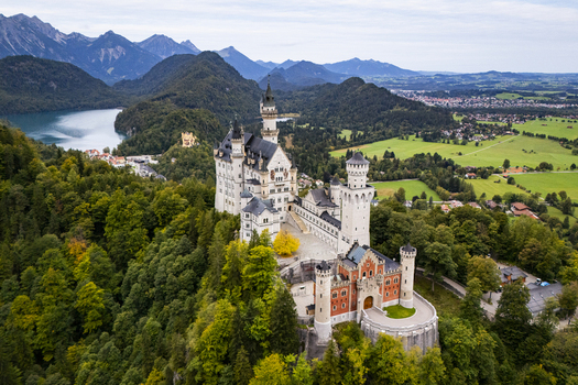 Aerial view of Neuschwanstein Castle rising majestically amidst vibrant green forests and serene blue waters framed by distant mountains Neuschwanstein Bayern Germany. Aerial view of Neuschwanstein Castle rising majestically amidst vibrant green forests and serene blue waters framed by distant mountains Neuschwanstein Bayern Germany.