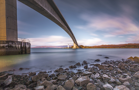 View of Skye Bridge spanning the water with a rocky shoreline in the foreground under a soft sunset sky in Isle of Skye United Kingdom. View of Skye Bridge spanning the water with a rocky shoreline in the foreground under a soft sunset sky in Isle of Skye United Kingdom.
