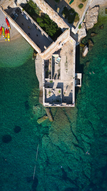 Aerial view of the historic stone fortress ruins on the turquoise coastline with colorful kayaks and swimmers in the clear water in Komiza Split Dalmatia County Croatia.