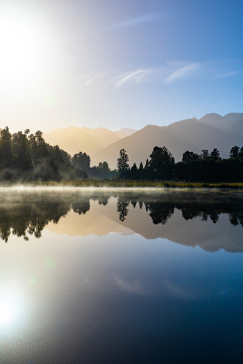 View of the still lake mirroring the silhouette of distant mountains under a soft hazy sky creating a serene and ethereal landscape Lake Matheson New Zealand. View of the still lake mirroring the silhouette of distant mountains under a soft hazy sky creating a serene and ethereal landscape Lake Matheson New Zealand.