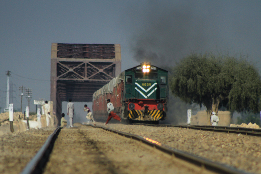 View of a freight train approaching a large steel bridge in the Cholistan Desert with people walking along the tracks in Bahawalpur Punjab Pakistan. View of a freight train approaching a large steel bridge in the Cholistan Desert with people walking along the tracks in Bahawalpur Punjab Pakistan.