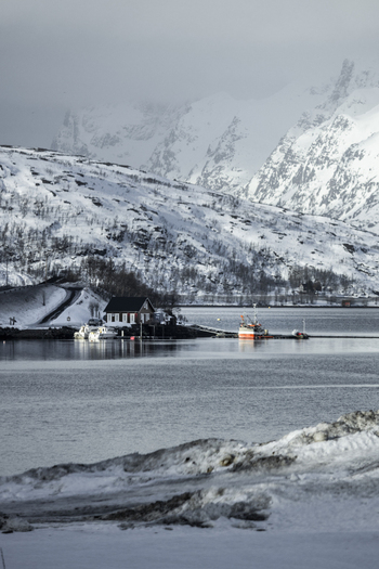 View of a small red cabin and fishing boats on the calm water with snow covered mountains under a cloudy sky in Kvaloya Troms Norway. View of a small red cabin and fishing boats on the calm water with snow covered mountains under a cloudy sky in Kvaloya Troms Norway.