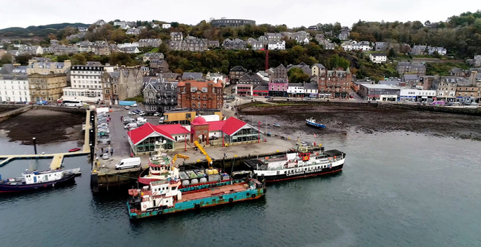 Oban United Kingdom 30 September 2026 Aerial view of Oban Ferry Terminal with ferries docked at the pier surrounded by the coastal town buildings hills and calm water under an overcast sky.