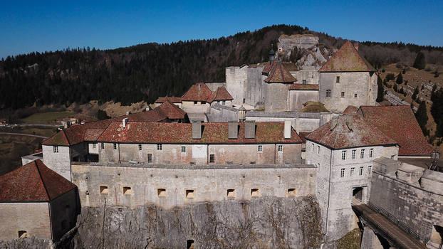 Aerial view of the historic Chateau de Joux fortress on a rocky outcrop with stone walls red tiled roofs and forested hills in La Cluse et Mijoux Bourgogne Franche Comte France. Aerial view of the historic Chateau de Joux fortress on a rocky outcrop with stone walls red tiled roofs and forested hills in La Cluse et Mijoux Bourgogne Franche Comte France.