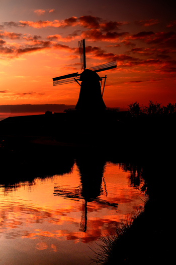 View of a traditional windmill silhouetted against a vibrant orange sunset sky with its reflection visible in the calm water below in the Netherlands. View of a traditional windmill silhouetted against a vibrant orange sunset sky with its reflection visible in the calm water below in the Netherlands.