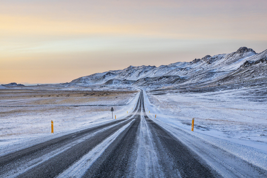 View of a stark asphalt road slicing through a frozen snow laden landscape framed by distant mountains under a pale sunrise sky Kleifarvatn Grindav&iacute;kurb&aelig;r Iceland.