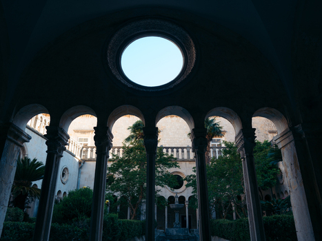 View of the stone arches and oculus of a historic cloister overlooking a lush courtyard with a fountain and palm trees in Dubrovnik Dubrovnik Neretva County Croatia. View of the stone arches and oculus of a historic cloister overlooking a lush courtyard with a fountain and palm trees in Dubrovnik Dubrovnik Neretva County Croatia.