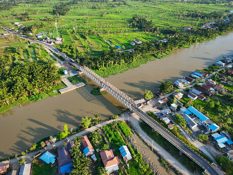 Aerial view of a steel truss bridge spanning a wide river surrounded by vibrant green rice paddies tropical palm trees and residential houses in Banjar Regency South Kalimantan Indonesia. Aerial view of a steel truss bridge spanning a wide river surrounded by vibrant green rice paddies tropical palm trees and residential houses in Banjar Regency South Kalimantan Indonesia.