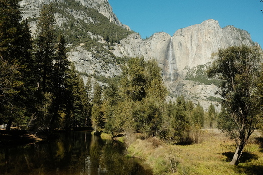 View of Yosemite Falls cascading down granite cliffs with the Merced River and lush green trees in the foreground California United States.