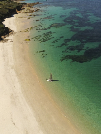 Aerial view of a small catamaran sailing in the clear turquoise waters off the sandy beach of Ile de Groix Groix Bretagne France. Aerial view of a small catamaran sailing in the clear turquoise waters off the sandy beach of Ile de Groix Groix Bretagne France.