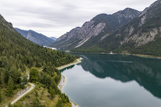 Aerial view of the serene Plansee lake reflecting the sky and the surrounding mountains with a winding road cutting through the lush green forests Plansee Tyrol Austria. Aerial view of the serene Plansee lake reflecting the sky and the surrounding mountains with a winding road cutting through the lush green forests Plansee Tyrol Austria.