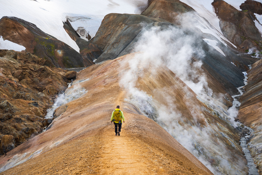 View of a hiker with a yellow backpack walking along a geothermal ridge in Kerlingarfjoll with rising steam volcanic soil and snow capped mountains in Hrunamannahreppur Iceland.