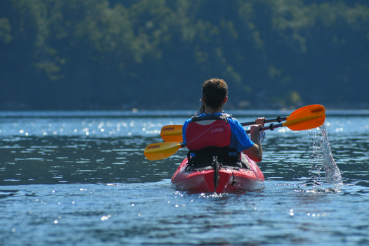 View of a person paddling a red kayak with orange oars on the calm blue waters of Nahuel Huapi Lake with lush green forests in the background San Carlos de Bariloche Rio Negro Argentina. View of a person paddling a red kayak with orange oars on the calm blue waters of Nahuel Huapi Lake with lush green forests in the background San Carlos de Bariloche Rio Negro Argentina.