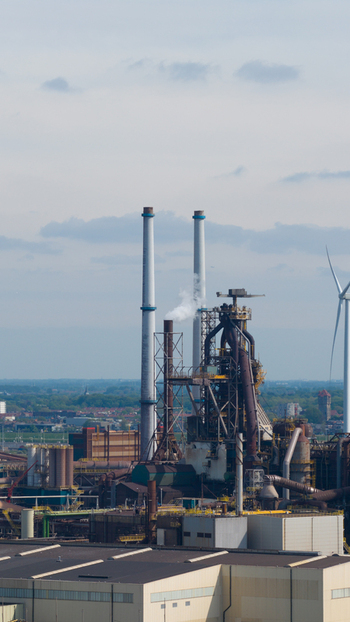 Aerial view of Tata Steel industrial plant featuring blast furnaces tall chimneys emitting steam and a wind turbine under a cloudy sky in Velsen Noord Noord Holland Netherlands. Aerial view of Tata Steel industrial plant featuring blast furnaces tall chimneys emitting steam and a wind turbine under a cloudy sky in Velsen Noord Noord Holland Netherlands.