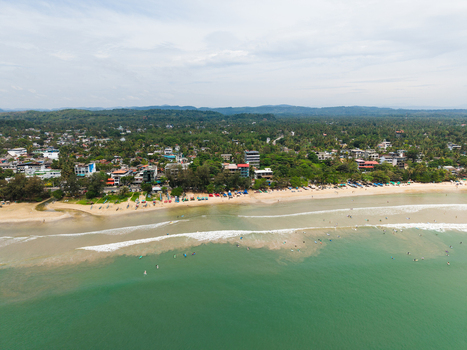 Aerial view of surfers in turquoise water along the sandy coastline and lush tropical trees in Weligama Southern Province Sri Lanka.