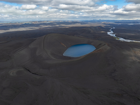 Aerial view of a volcanic crater lake with deep blue water surrounded by dark volcanic terrain and vast lava fields under a cloudy sky in Iceland. Aerial view of a volcanic crater lake with deep blue water surrounded by dark volcanic terrain and vast lava fields under a cloudy sky in Iceland.
