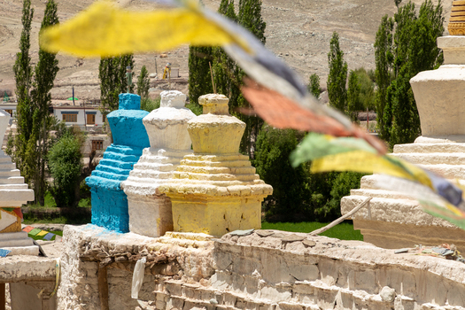 View of Charasa Gonpa featuring colorful stupas in blue white and yellow with prayer flags in the foreground against arid mountains and green trees in Charasa Ladakh India.