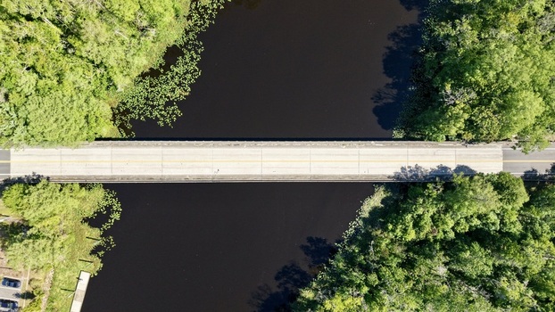 Aerial view of Sixmile Creek bridge crossing dark water surrounded by dense green forest and lily pads in St. Johns County Florida United States.