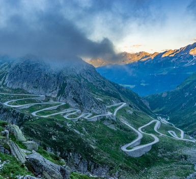 View of the winding Sankt Gotthard Pass road snaking through the rugged alpine landscape with dramatic clouds and sunset light Airolo Ticino Switzerland.