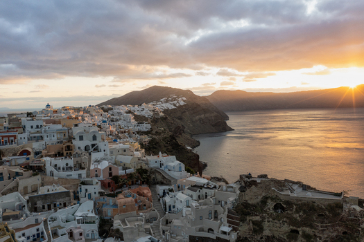 Aerial view of white buildings cascade down cliffs towards the turquoise sea as the sun sets behind the hills Fira Thira Greece. Aerial view of white buildings cascade down cliffs towards the turquoise sea as the sun sets behind the hills Fira Thira Greece.