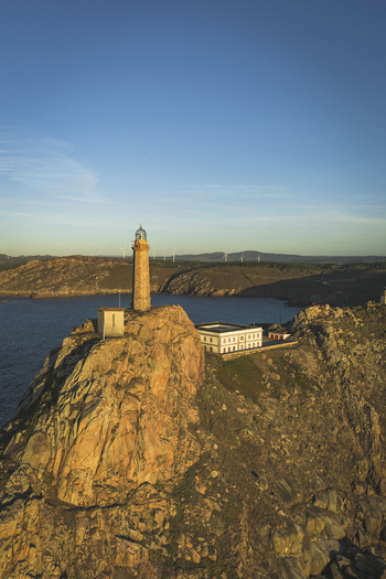 Aerial view of the lighthouse perched on a rocky cliff overlooking the Atlantic Ocean with wind turbines in the distance in Galicia Spain.