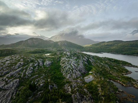Aerial view of the rugged rocky terrain featuring green vegetation small pools of water and mountains shrouded in clouds under soft light in Lofoten Norway.