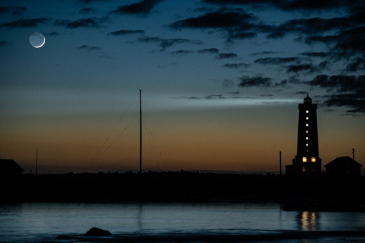 View of Grotta Lighthouse silhouetted against a twilight sky with a crescent moon and dark clouds over the water in Seltjarnarnes Seltjarnarnesbaer Iceland. View of Grotta Lighthouse silhouetted against a twilight sky with a crescent moon and dark clouds over the water in Seltjarnarnes Seltjarnarnesbaer Iceland.