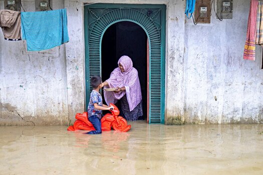 Banshkhali Upazila Bangladesh 10 July 2026 View of a woman in a hijab handing food to a young boy sitting on a red toy motorcycle in floodwaters in front of their home. Banshkhali Upazila Bangladesh 10 July 2026 View of a woman in a hijab handing food to a young boy sitting on a red toy motorcycle in floodwaters in front of their home.