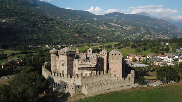 Aerial view of the medieval Fenis Castle featuring stone towers and crenelated walls set against a backdrop of mountains and green fields in Aosta Valley Italy. Aerial view of the medieval Fenis Castle featuring stone towers and crenelated walls set against a backdrop of mountains and green fields in Aosta Valley Italy.