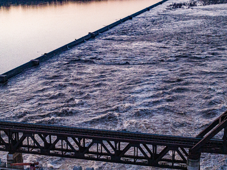 Aerial view of turbulent river water rushing over a dam structure near a steel truss bridge during sunset in Louisville Kentucky United States.