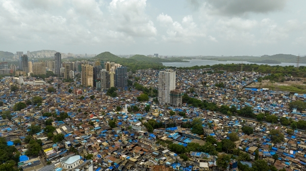 Aerial view of Dharavi Slums with dense residential rooftops modern high rise buildings lush hills and a distant water body under a cloudy sky in Mumbai Maharashtra India. Aerial view of Dharavi Slums with dense residential rooftops modern high rise buildings lush hills and a distant water body under a cloudy sky in Mumbai Maharashtra India.